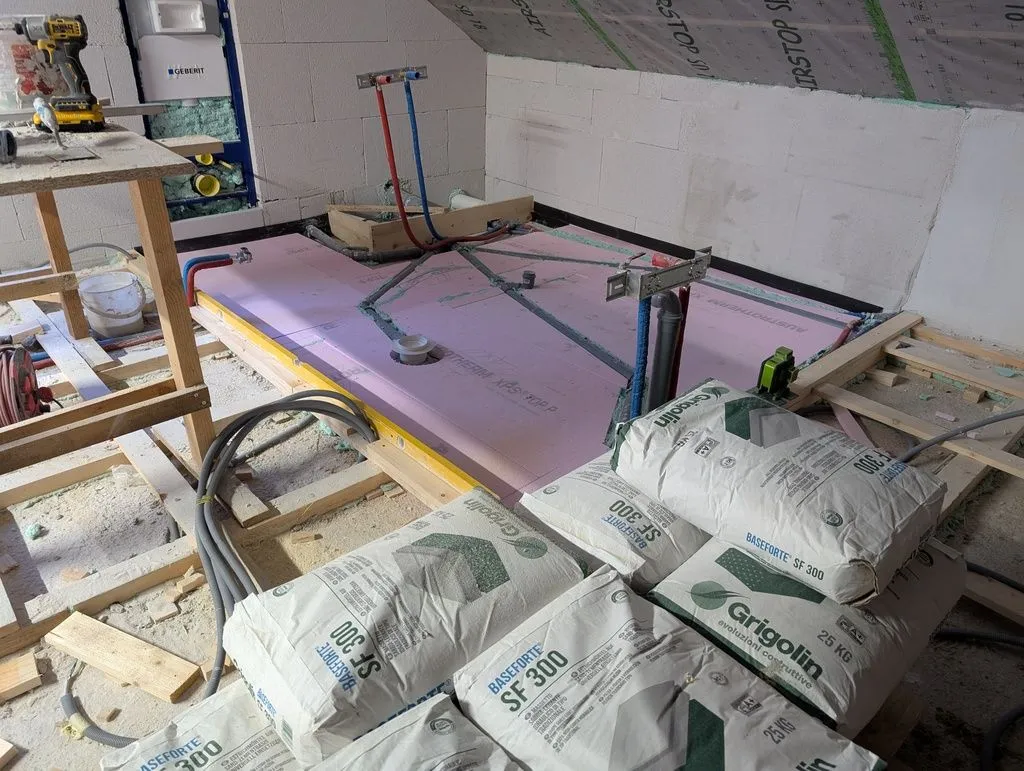 We first placed 50 mm XPS over the floor (we use a bottom layer of XPS instead of screed all the way down because it lessens the dead load on the ceiling below, and because it is cheaper and easier to install). The black tape around the perimeter of the bathroom is a foam-like material that serves as an isolation joint for the screed. 